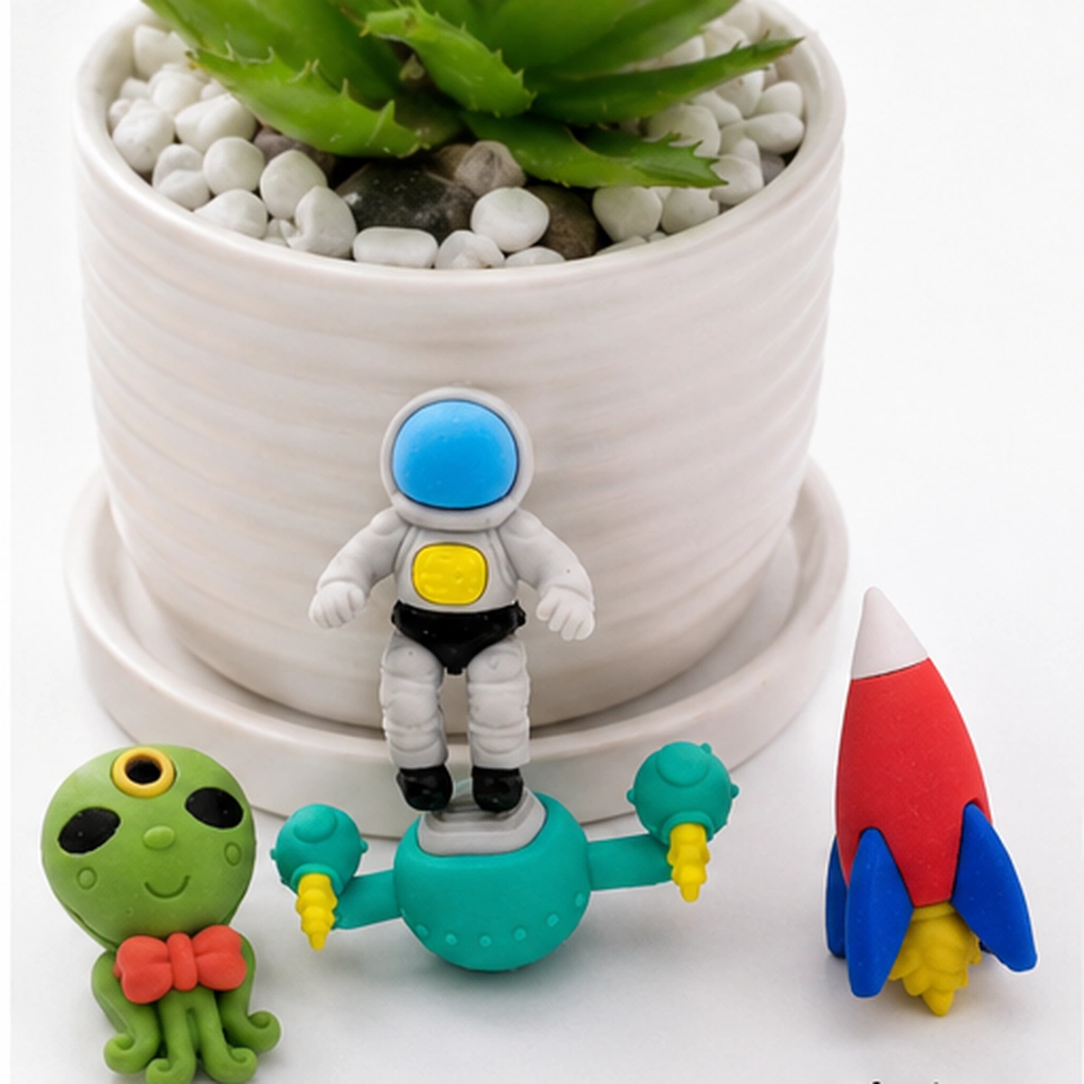Astronaut eraser standing on the teal spacecraft beside the alien octopus and red rocket, in front of a white plant pot